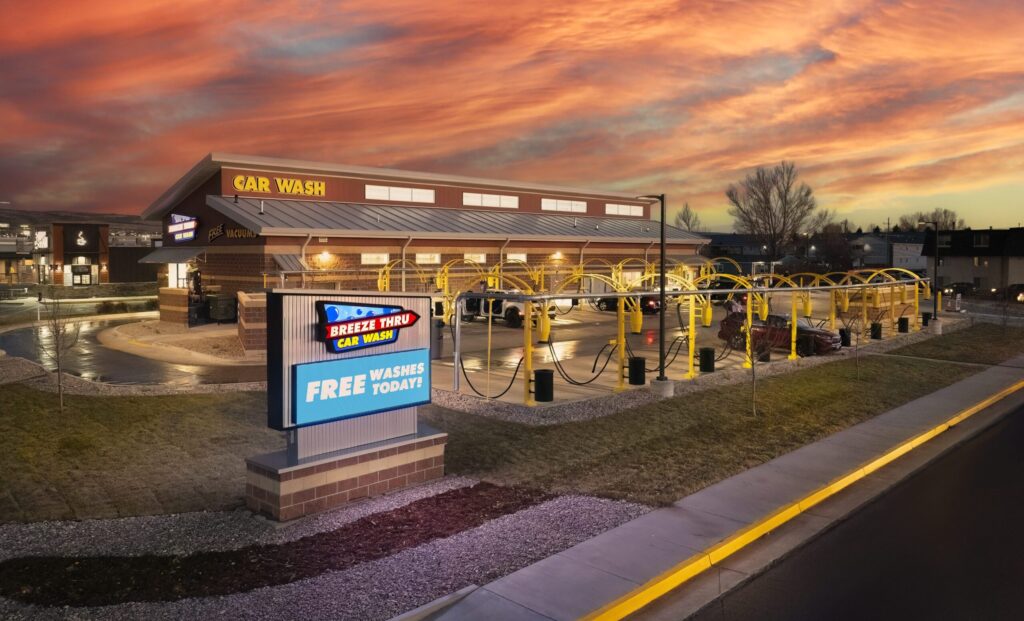 A wide view of Breeze Thru Car Wash in Laramie, WY at dusk, with an LED message center and illuminated building signage glowing against a bright orange and pink sunset.
shine