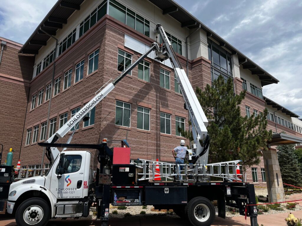 Technicians installing a building-mounted sign using a Schlosser Signs bucket truck on a multi-story commercial building. Permitting