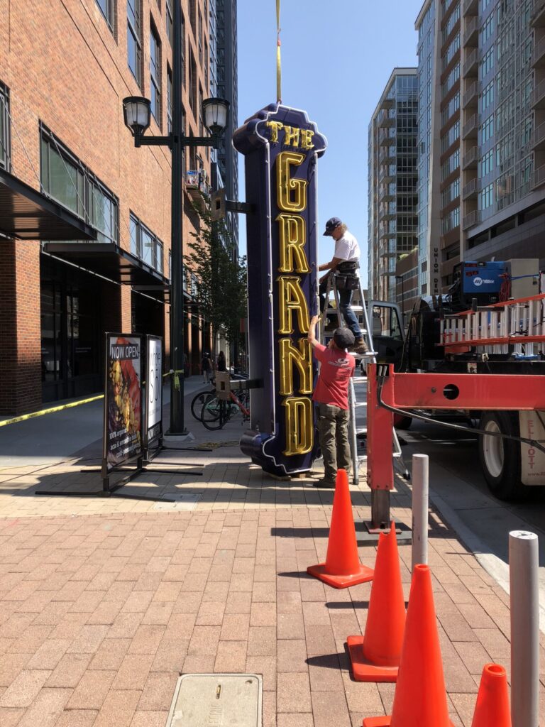 Crew installing a large illuminated “The Grand” vertical sign on a city sidewalk between multi-story buildings, with equipment and safety cones in place. Permitting
